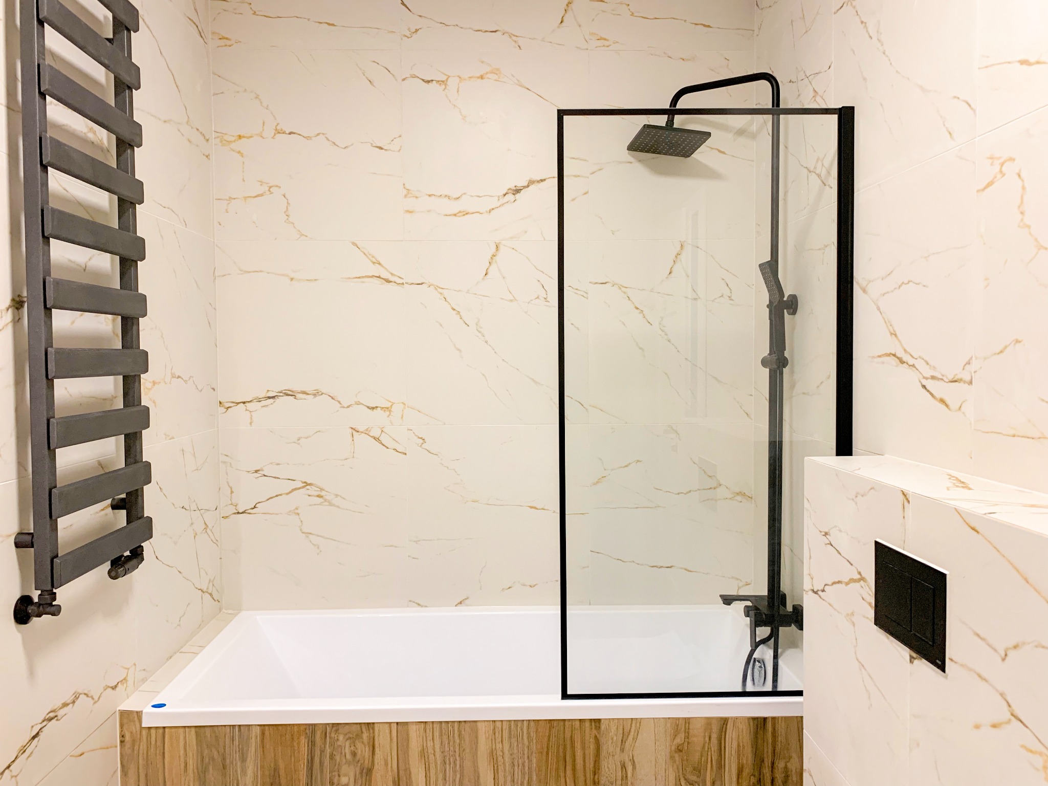 Wykończona łazienka z wanną obudowaną drewnopodobnymi panelami, czarną armaturą i grzejnikiem drabinkowym na tle płytek imitujących marmur z złotymi żyłami.