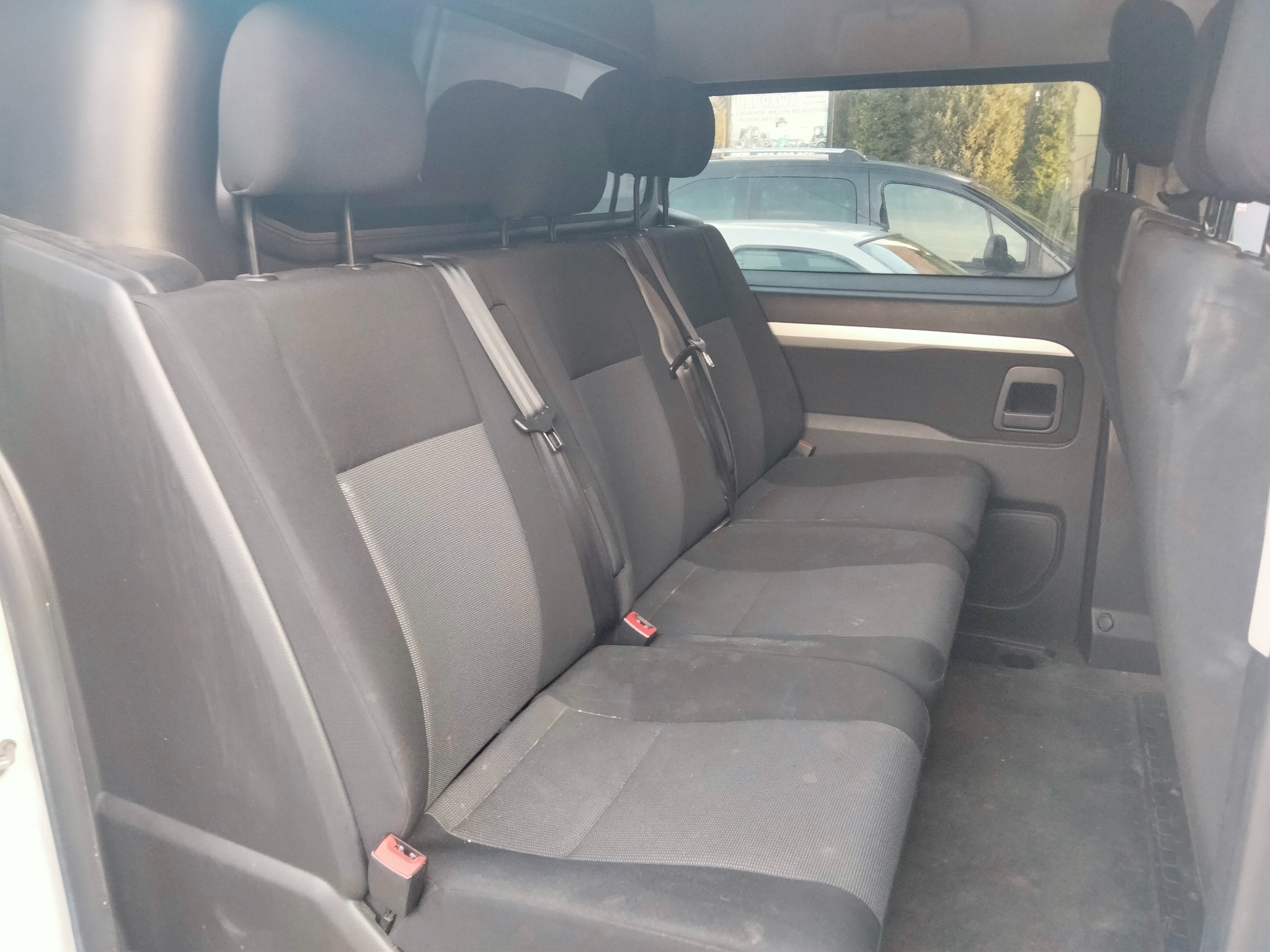 Wnętrze pojazdu dostawczego z trzema siedzeniami z tapicerką w odcieniach szarości przed czyszczeniem, widoczne plamy i zabrudzenia na siedzeniach i podłodze.