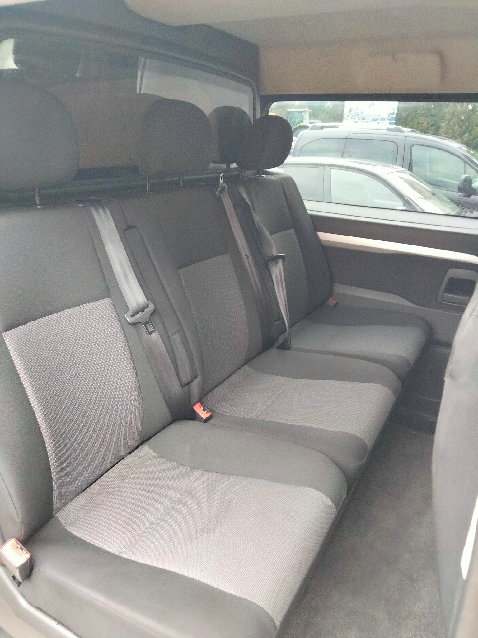 Wnętrze busa z trzema rzędami szarych foteli tapicerowanych, widoczne pasy bezpieczeństwa i fragmenty innych pojazdów za oknem.