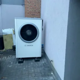 Biała pompa ciepła Bosch ustawiona na czarnych podstawkach przed budynkiem z ceglanym kominem w tle.