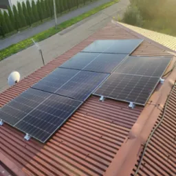 Panele słoneczne zamontowane na falistym, czerwonym dachu z widoczną anteną satelitarną i okoliczną zabudową w tle, uchwycone w słoneczny dzień.