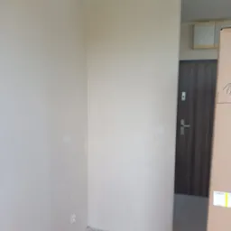 Wnętrze pokoju po nałożeniu gładzi, ściany w jasnym, neutralnym odcieniu, widoczna szara podłoga i fragment brązowych drzwi w tle.