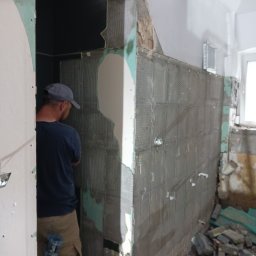 Raz a Dobrze. - Mężczyzna w czapce glazurniczej przykłada płytki do ściany w trakcie prac wykończeniowych w łazience, widoczne nierówne krawędzie i resztki starej glazury, w tle okno i gruzowisko.