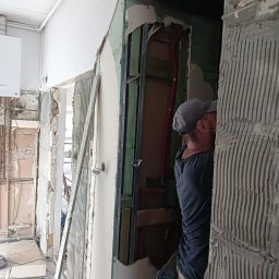 Raz a Dobrze. - Demontaż płytek w łazience podczas remontu, widoczne odsłonięte instalacje i fragment pracującego glazurnika w szarej czapce.