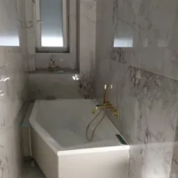 Biała wanna obudowana płytami gipsowo-kartonowymi, złota bateria łazienkowa, marmurowe płytki na ścianach, małe okno z parapetem.