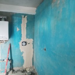 Raz a Dobrze. - Niebieska ściana w trakcie prac wykończeniowych. Widoczny fragment instalacji, poziomica i gruz na podłodze. Ściana częściowo pokryta gładzią.