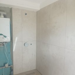 Raz a Dobrze. - Jasne, pionowe płytki w trakcie montażu w łazience w Dzierżoniowie. Widoczne przyłącza i odpływ liniowy. Częściowo wykończona ściana z piecykiem.