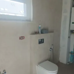 Podwieszana toaleta w jasnej łazience z oknem. Widoczne płytki imitujące kamień i elementy instalacji. Częściowo wykończona łazienka w trakcie prac.