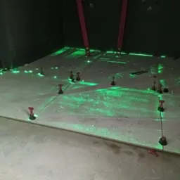 Układanie płytek na podłodze z użyciem poziomicy laserowej i systemu poziomowania płytek; widoczne krzyżyki dystansowe i poziomice pionowe przy ścianie w ciemnym pomieszczeniu.