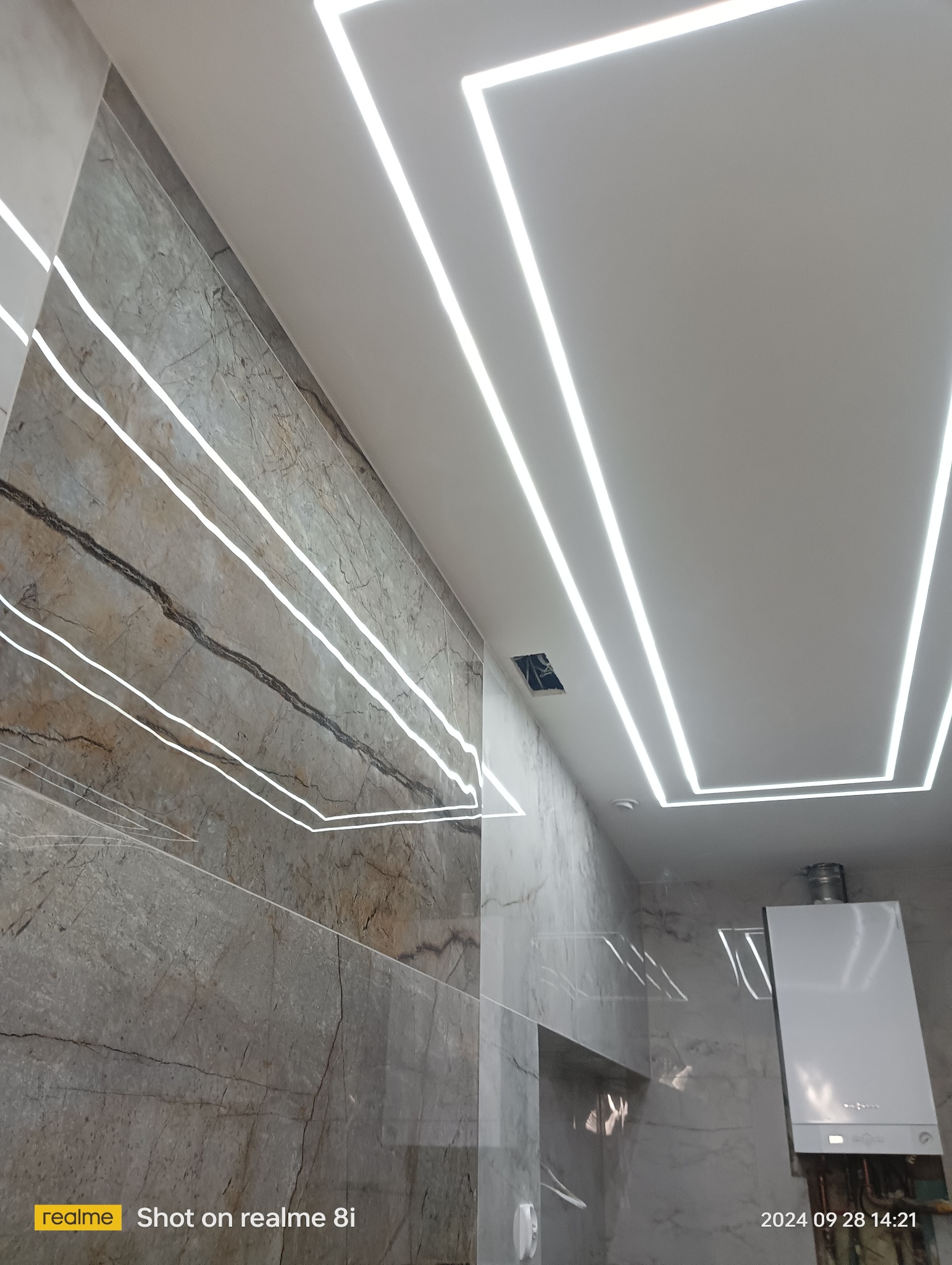 Wykończona łazienka z marmurowymi płytkami na ścianach, podświetlanym sufitem LED w kształcie prostokąta i zamontowanym piecem grzewczym.