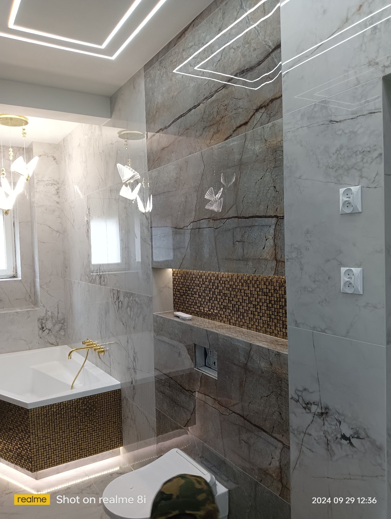 Nowoczesna łazienka z marmurowymi płytkami, złotą armaturą, podświetlanym sufitem i dekoracyjną mozaiką w niszy, z widoczną toaletą i wanną.