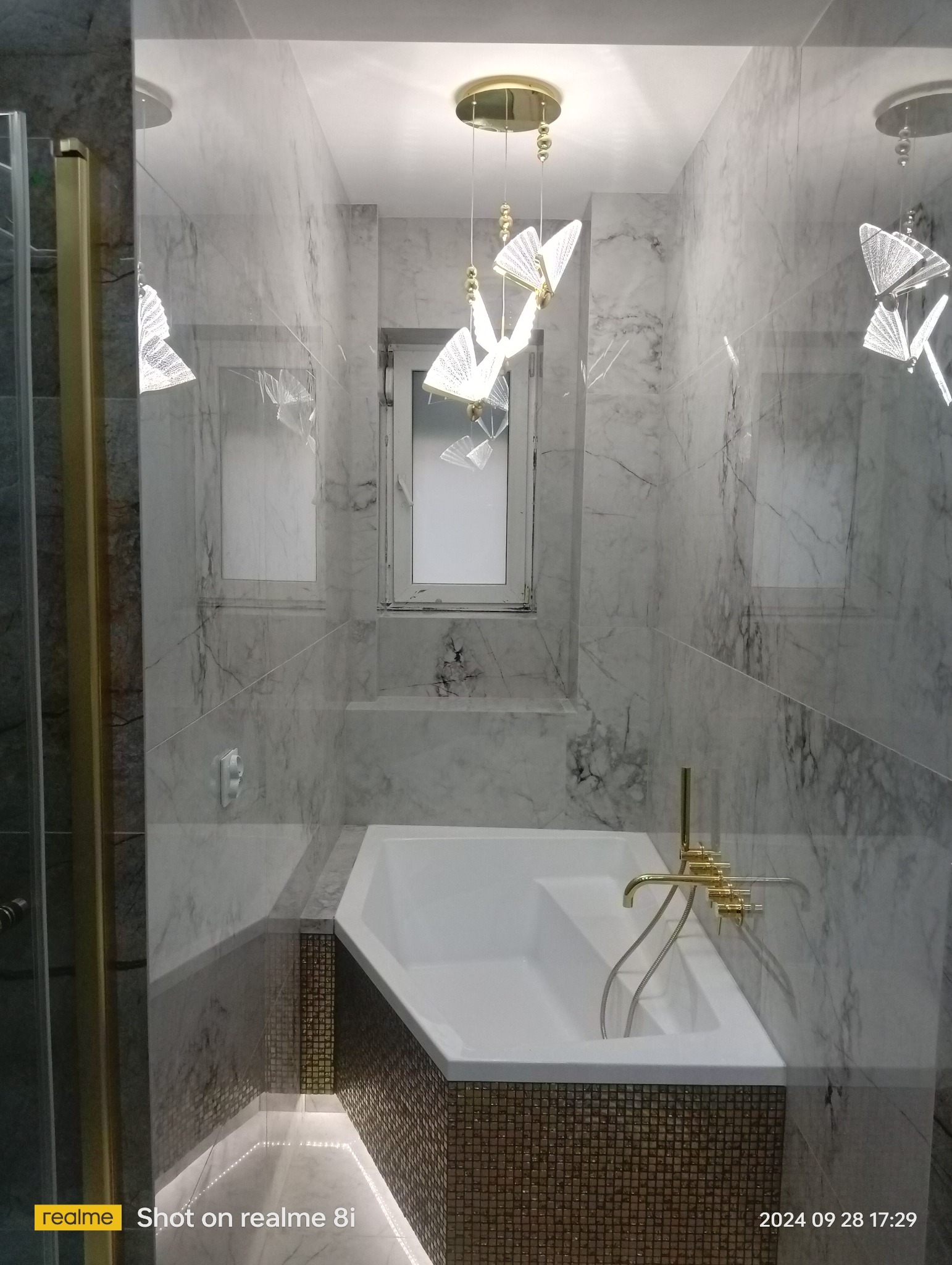 Nowoczesna łazienka z wanną obłożoną mozaiką, marmurowe ściany i podświetlane listwy LED pod wanną, oświetlenie sufitowe w kształcie motyli.