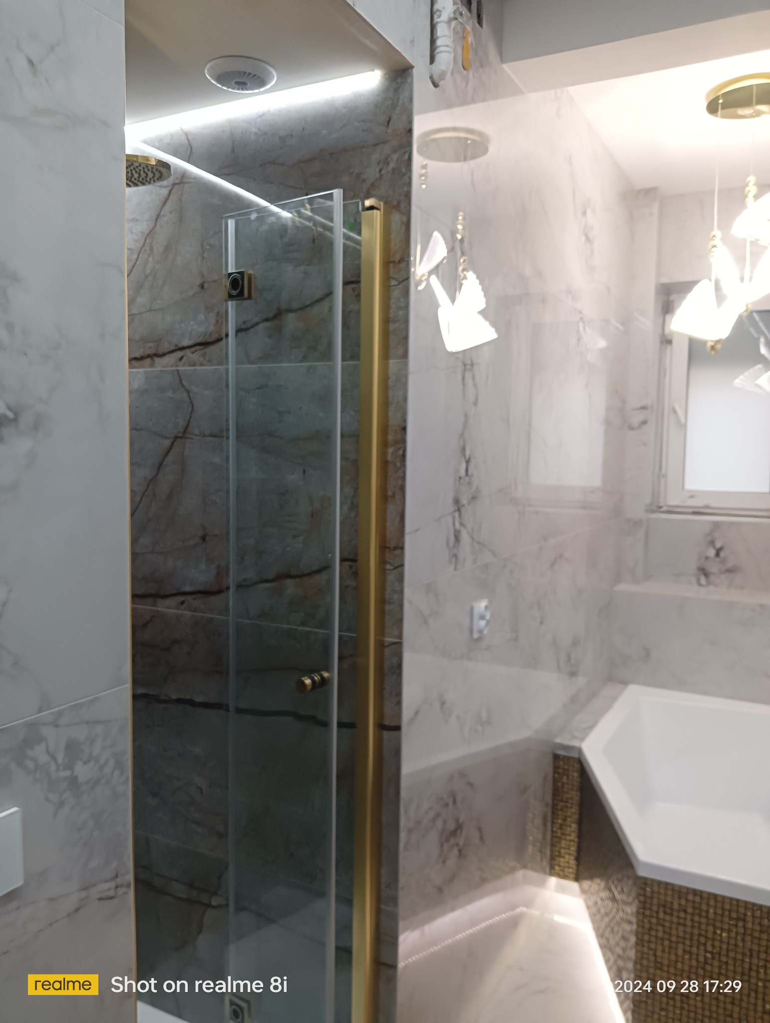 Szklana kabina prysznicowa z złotymi profilami na tle marmurowych płytek w nowoczesnej łazience z oświetleniem LED i wiszącą lampą.