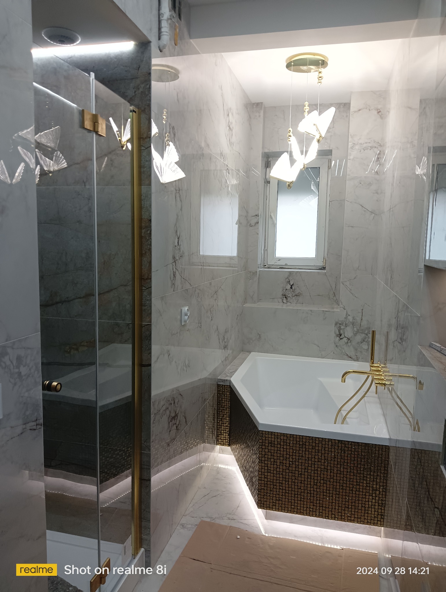 Nowoczesna łazienka z marmurowymi płytkami, kabiną prysznicową ze złotymi detalami i podświetlaną wanną z mozaiką, oświetlona designerską lampą.