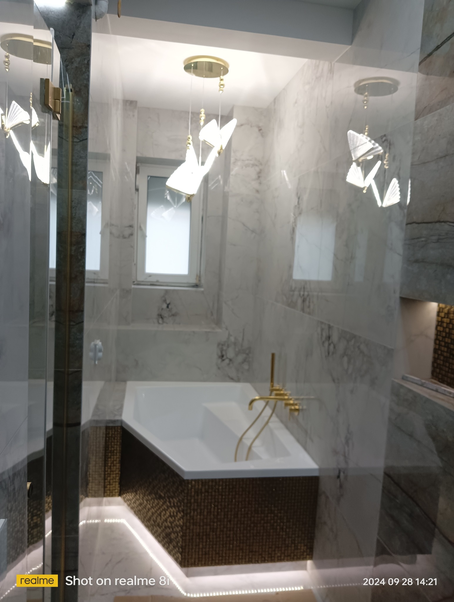 Nowoczesna łazienka z marmurowymi ścianami, podświetlaną wanną obłożoną mozaiką, złotą armaturą i designerskimi lampami w kształcie origami.