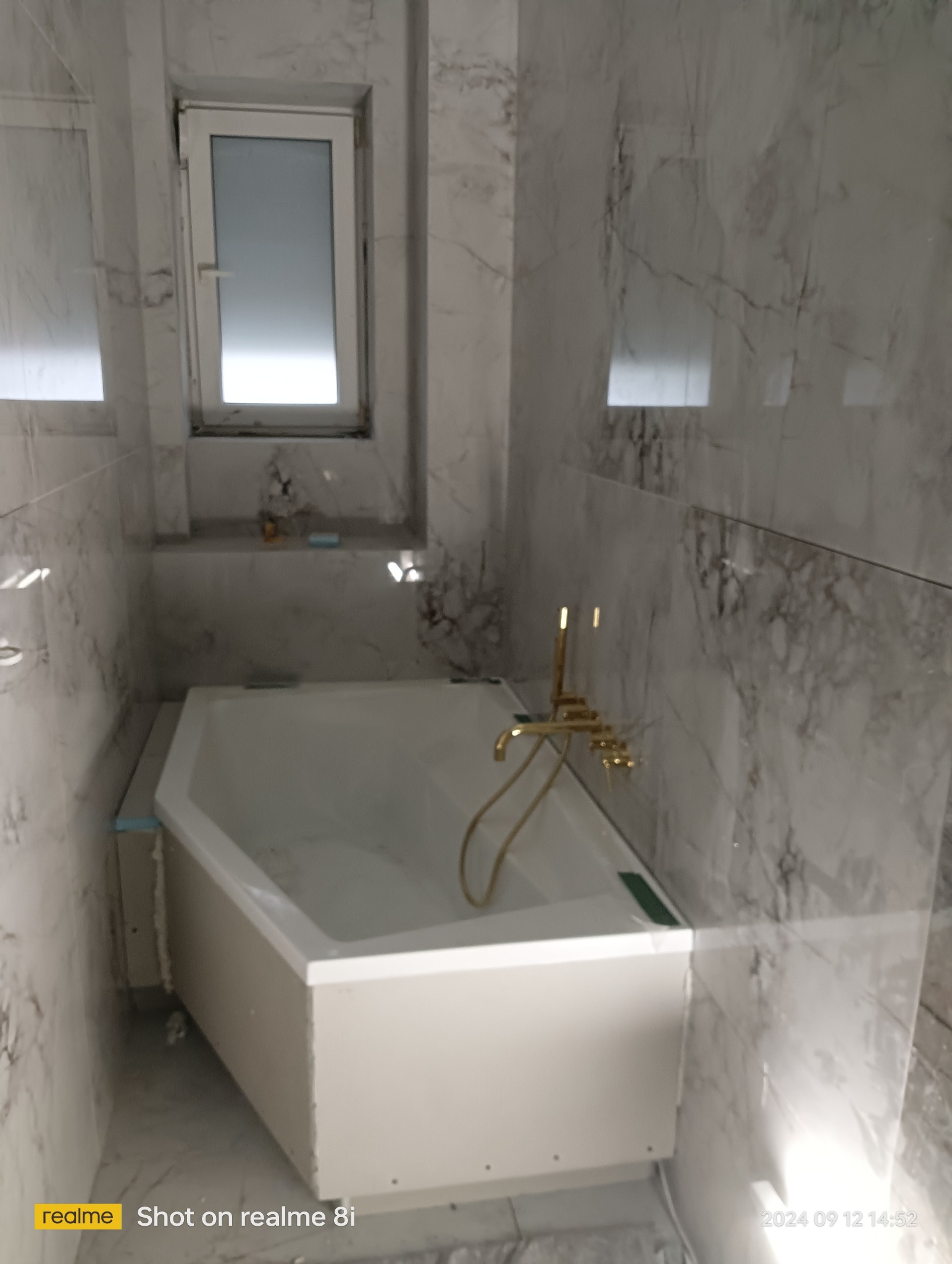 Biała wanna obudowana płytami gipsowo-kartonowymi, złota bateria łazienkowa, marmurowe płytki na ścianach, małe okno z parapetem.