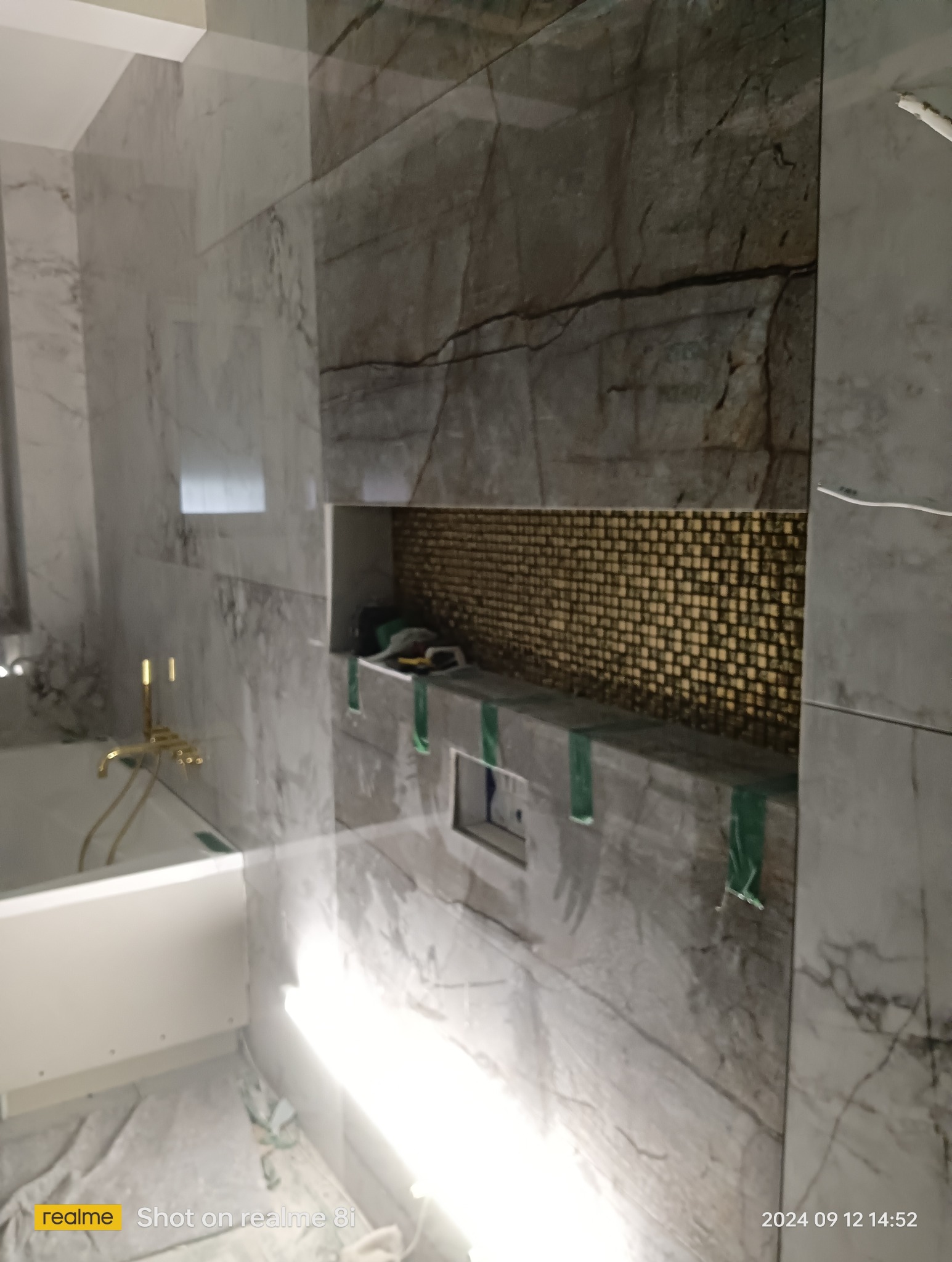 Wnęka w ścianie wyłożona mozaiką w łazience, widoczne zabezpieczenia taśmą malarską, bateria wannowa w kolorze złotym, marmurowe płytki na ścianach