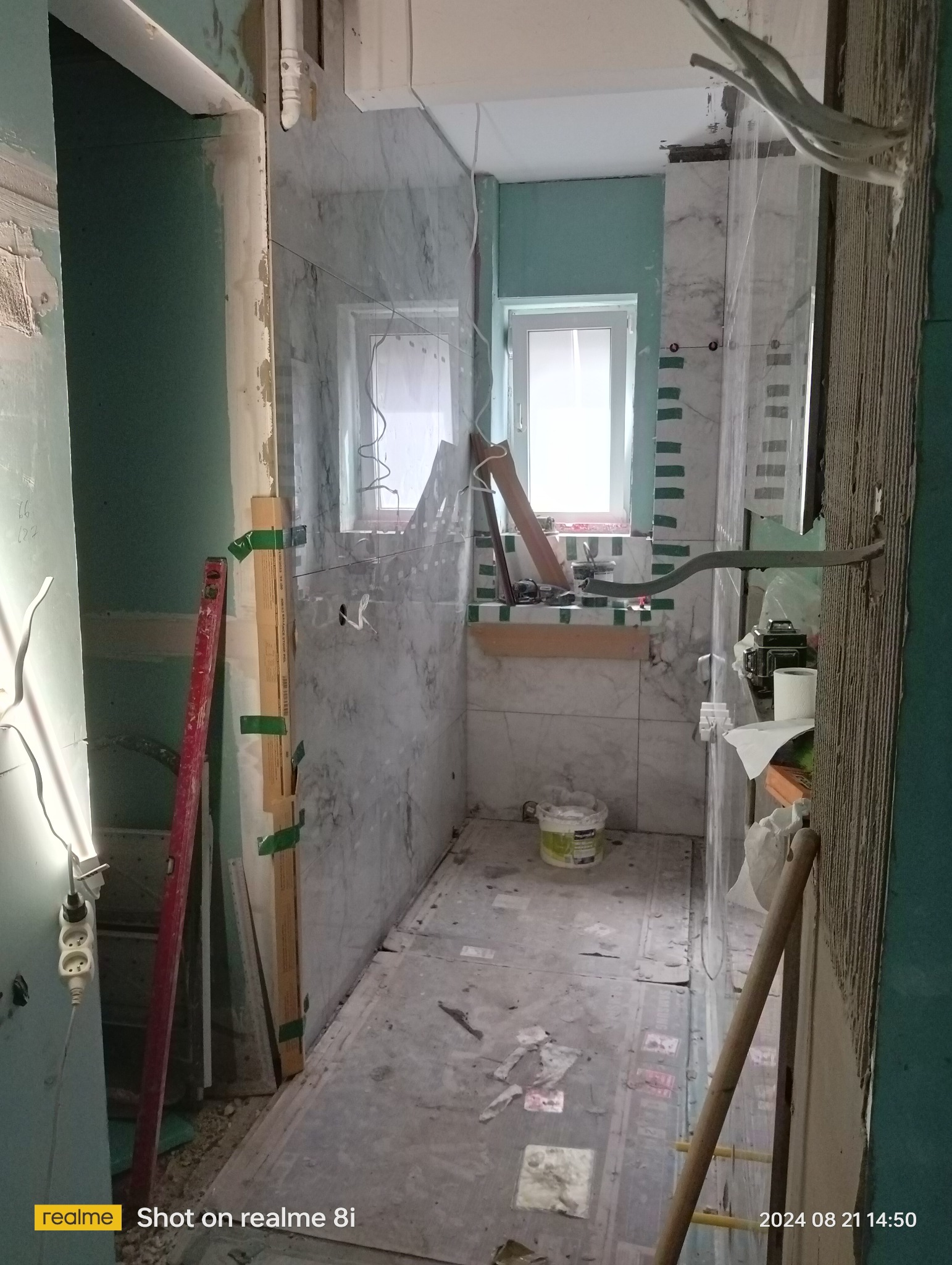Wnętrze łazienki w trakcie prac wykończeniowych, widoczne częściowo ułożone marmurowe płytki, okno z zieloną ramą, poziomica, przedłużacz i wiadro z zaprawą na podłodze.