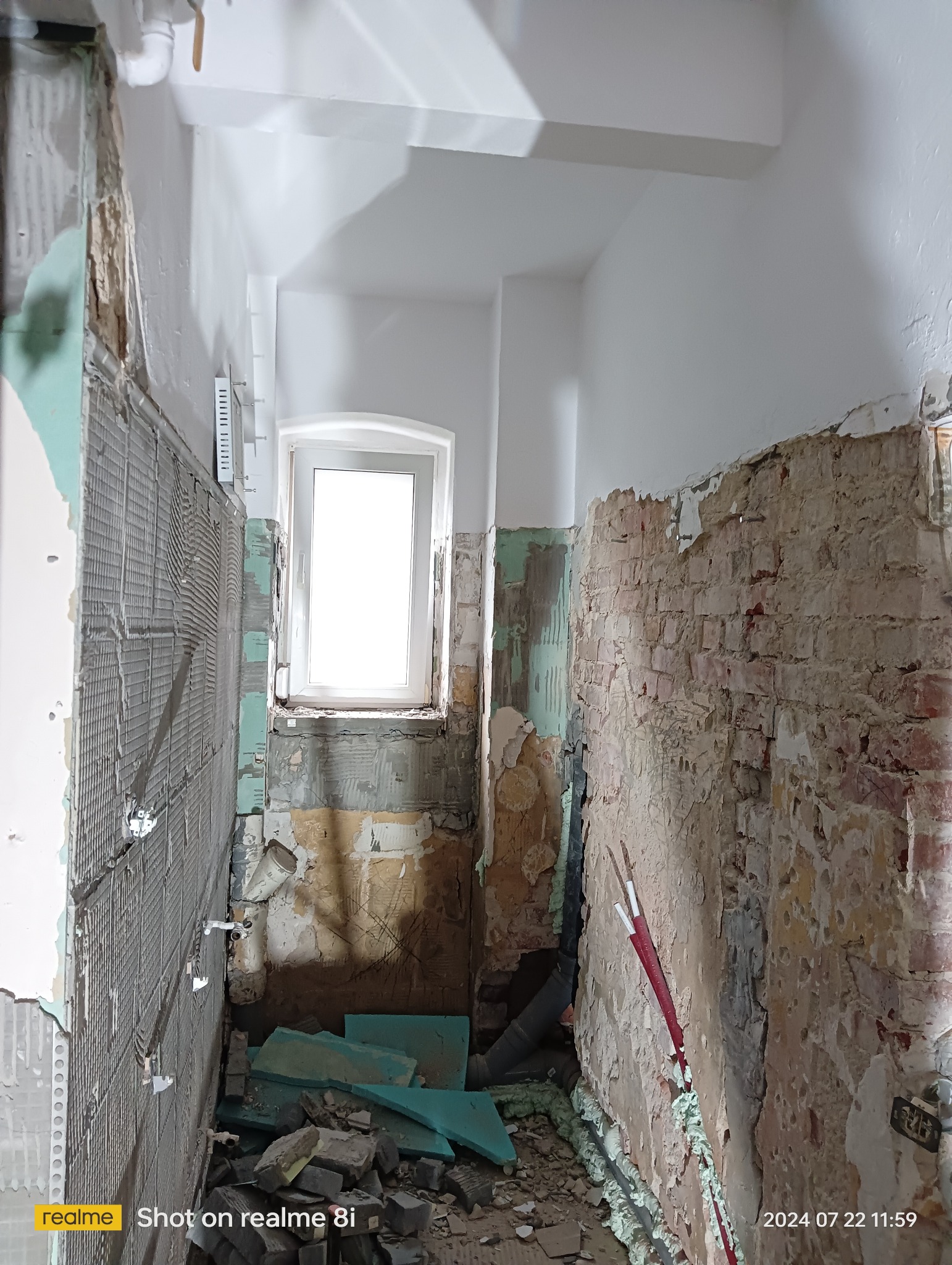 Wnętrze w trakcie demontażu starych płytek i przygotowania ścian pod nowe wykończenie. Widoczne fragmenty starej cegły, instalacje wodne i metalowa siatka na ścianie.