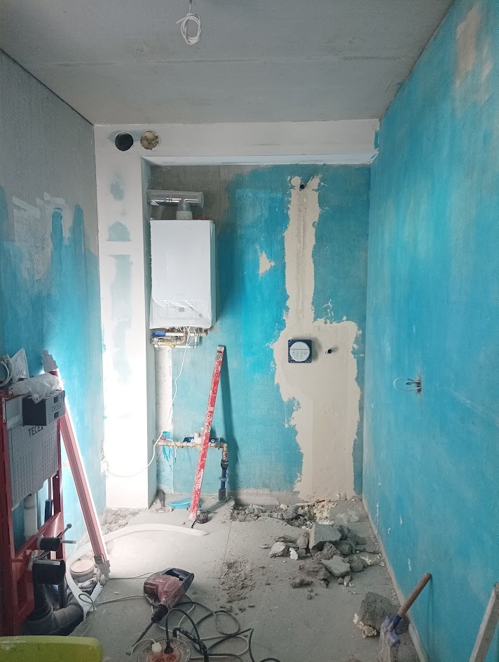 Wnętrze łazienki w trakcie prac: ściany w kolorze niebieskim i szarym, widoczny bojler, instalacja wodna, gruz na podłodze i narzędzia, w tym poziomica.