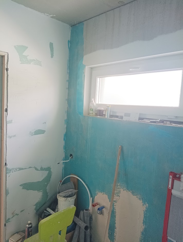 Narożnik łazienki w trakcie prac przygotowawczych: ściany częściowo pokryte gruntem, widoczne rury i narzędzia, okno z parapetem. Etap prac przed wykończeniem.