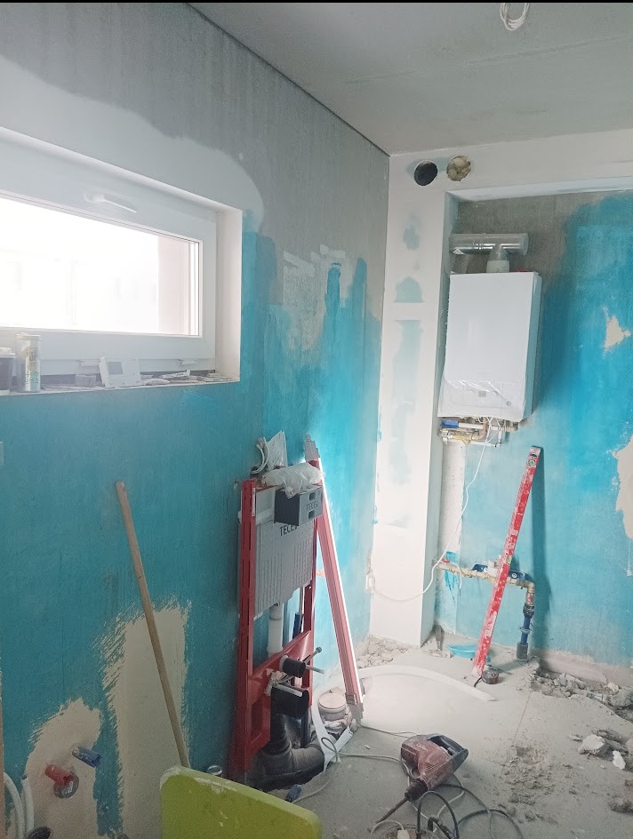 Wnętrze łazienki w trakcie prac: niebieskie ściany, częściowo położona gładź, stelaż WC, piec gazowy, poziomica i narzędzia. Widoczne ślady po kuciu.