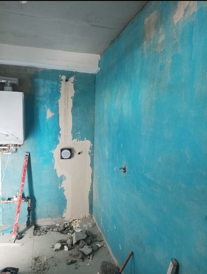 Niebieska ściana w trakcie prac wykończeniowych. Widoczny fragment instalacji, poziomica i gruz na podłodze. Ściana częściowo pokryta gładzią.