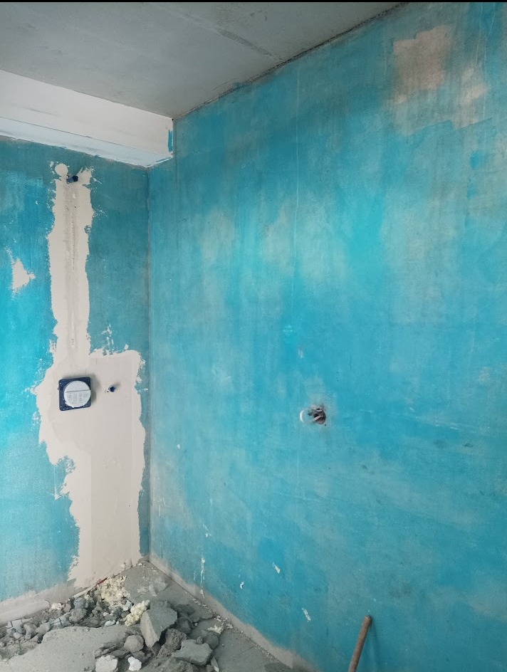 Niebieska ściana w trakcie remontu, częściowo zagruntowana, z widocznymi instalacjami i gruzem na podłodze. Przygotowanie do wykończenia wnętrza.