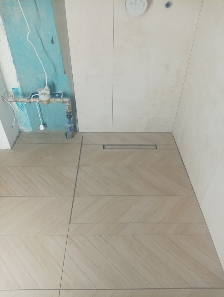 Ułożone beżowe płytki podłogowe w jodełkę z odpływem liniowym w łazience, widoczne fragmenty instalacji wodnej i surowej ściany.