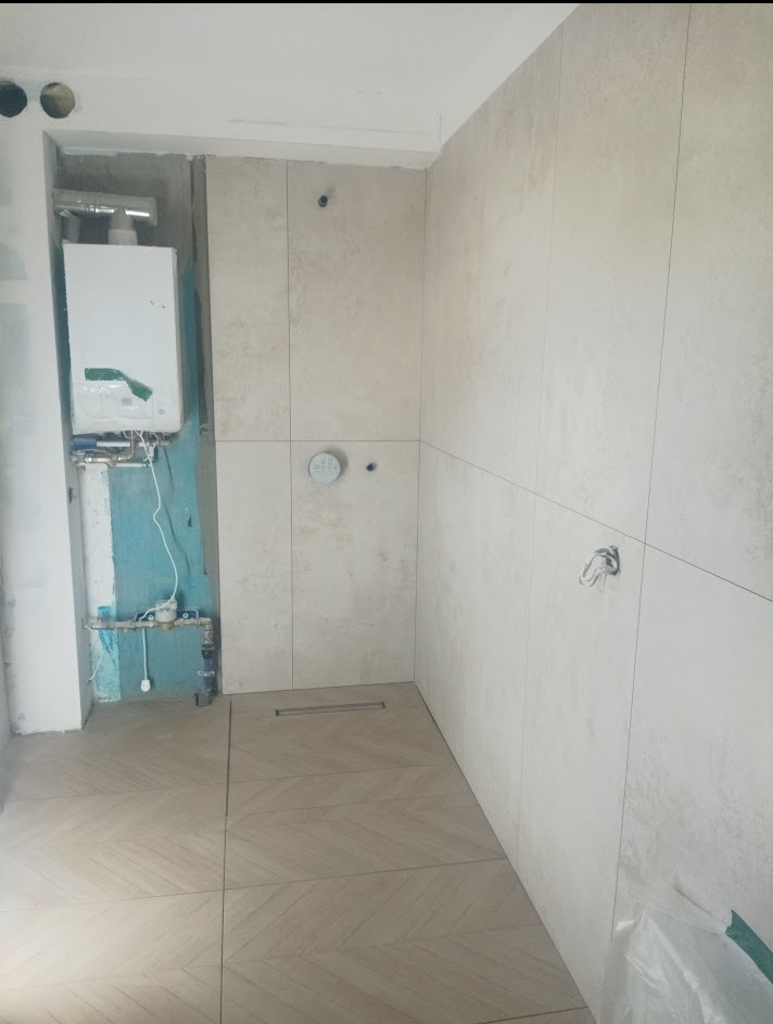 Jasne płytki na ścianach i podłodze w łazience w trakcie remontu. Widoczny bojler, odpływ liniowy i przygotowane otwory na armaturę.