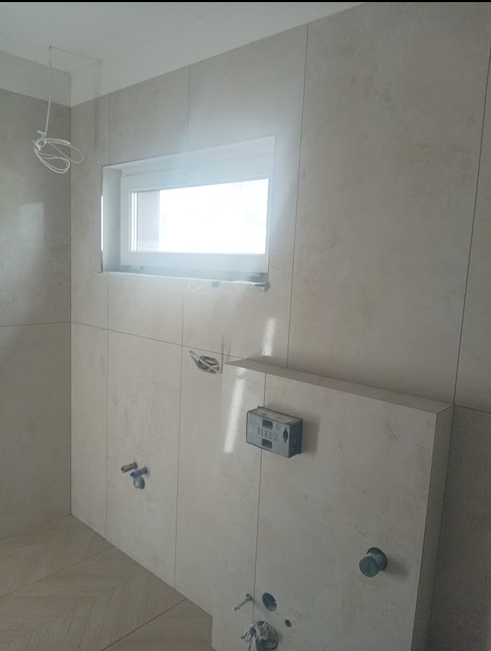 Łazienka w trakcie wykańczania: beżowe płytki, okno, instalacja sanitarna i elektryczna. Widoczne przygotowania do montażu armatury.