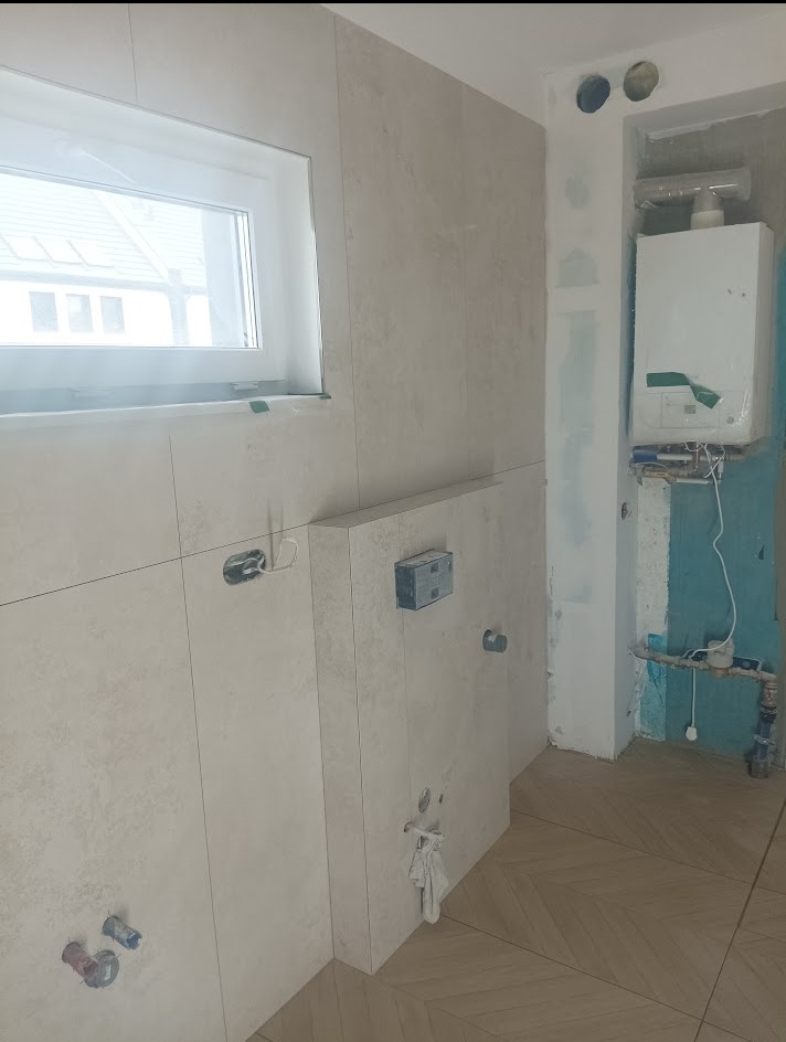 Beżowe płytki na ścianie w łazience w trakcie prac wykończeniowych, widoczne instalacje i piec gazowy. Parkiet ułożony w jodełkę.