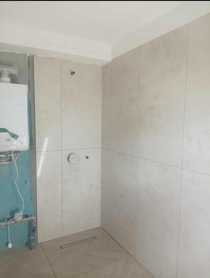Jasne, pionowe płytki w trakcie montażu w łazience w Dzierżoniowie. Widoczne przyłącza i odpływ liniowy. Częściowo wykończona ściana z piecykiem.