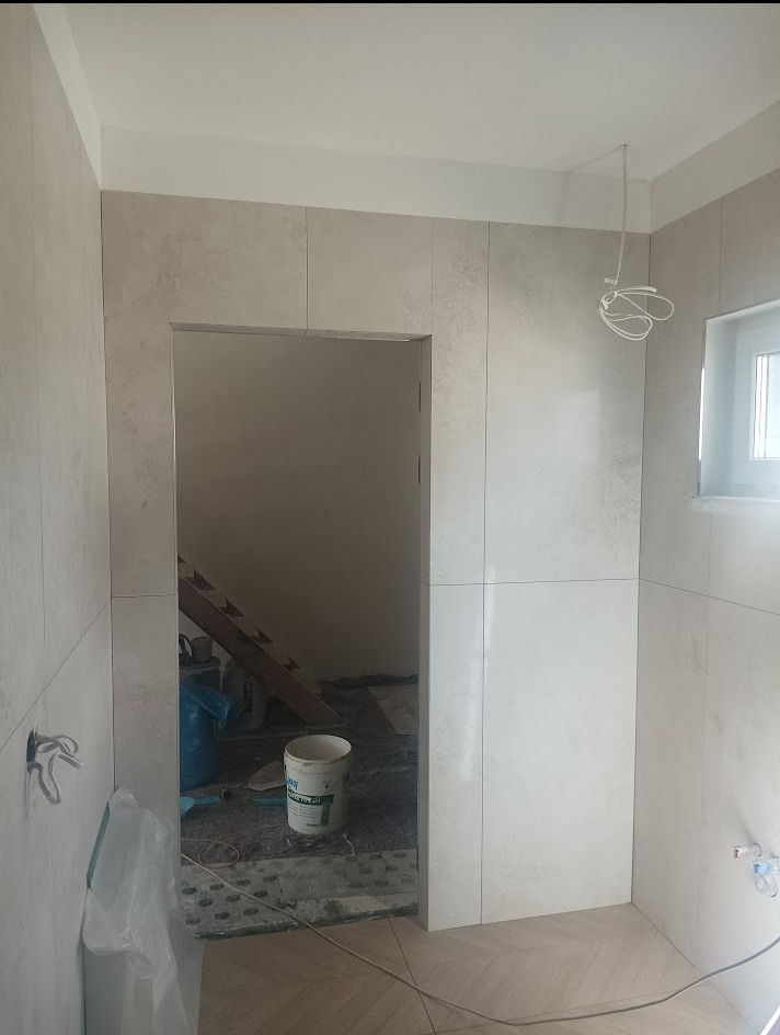 Wnętrze łazienki w trakcie prac wykończeniowych. Widoczne płytki na ścianach i podłodze, wiadro z zaprawą, przewody elektryczne. Fragment schodów w tle.