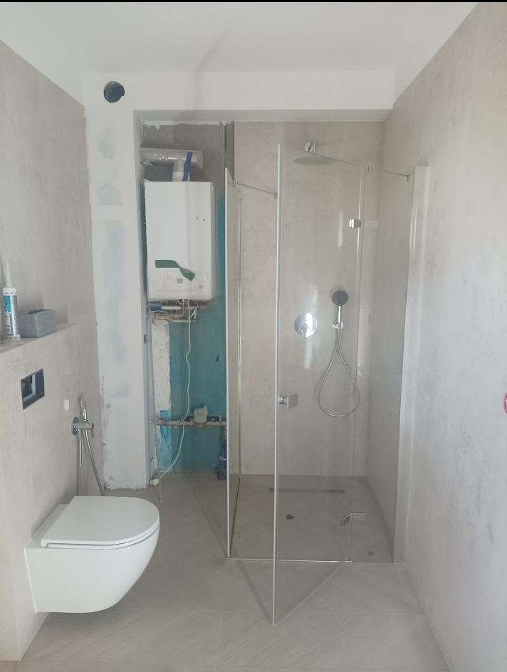 Wnętrze łazienki w trakcie remontu: kabina prysznicowa ze szklanymi drzwiami, podwieszana toaleta i piec gazowy na ścianie z widocznymi instalacjami.