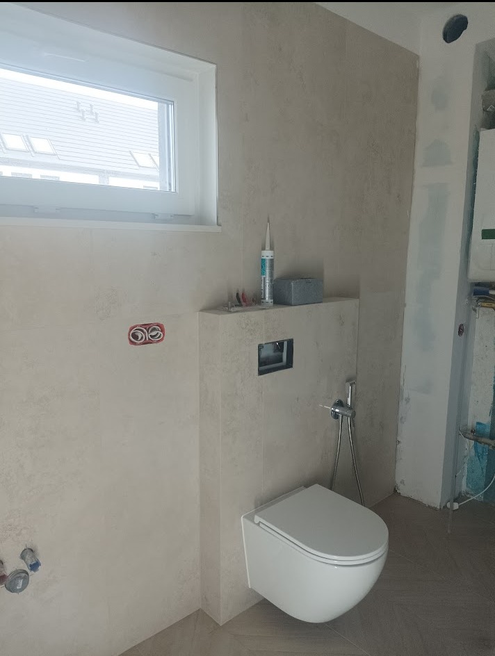 Podwieszana toaleta w jasnej łazience z oknem. Widoczne płytki imitujące kamień i elementy instalacji. Częściowo wykończona łazienka w trakcie prac.