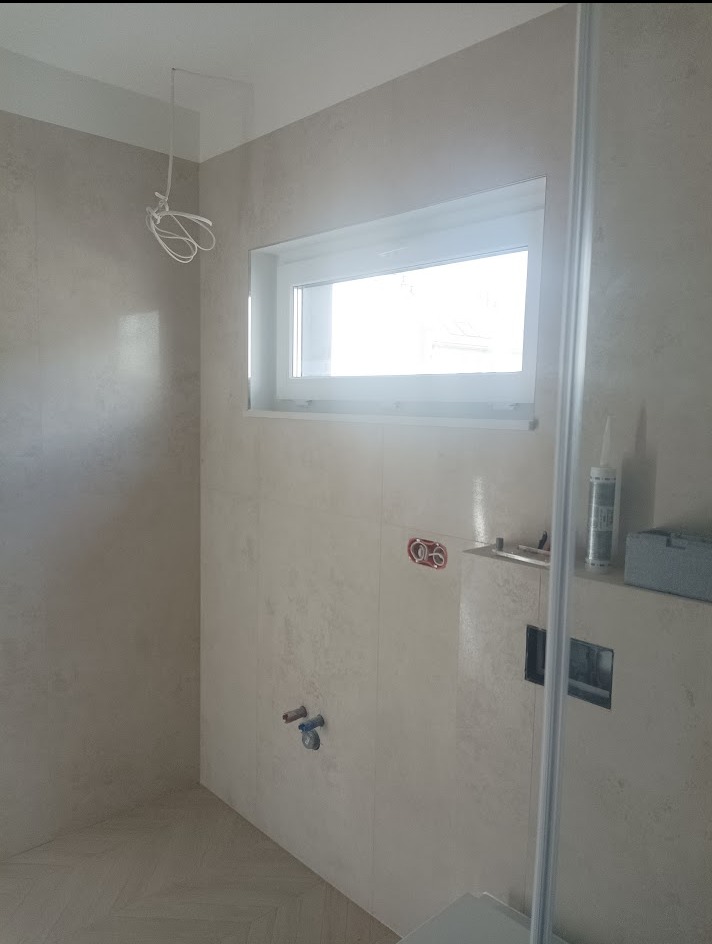Jasna łazienka w trakcie wykańczania: beżowe płytki, okno, wystające kable i rury. Widoczne materiały budowlane i fragment kabiny prysznicowej.