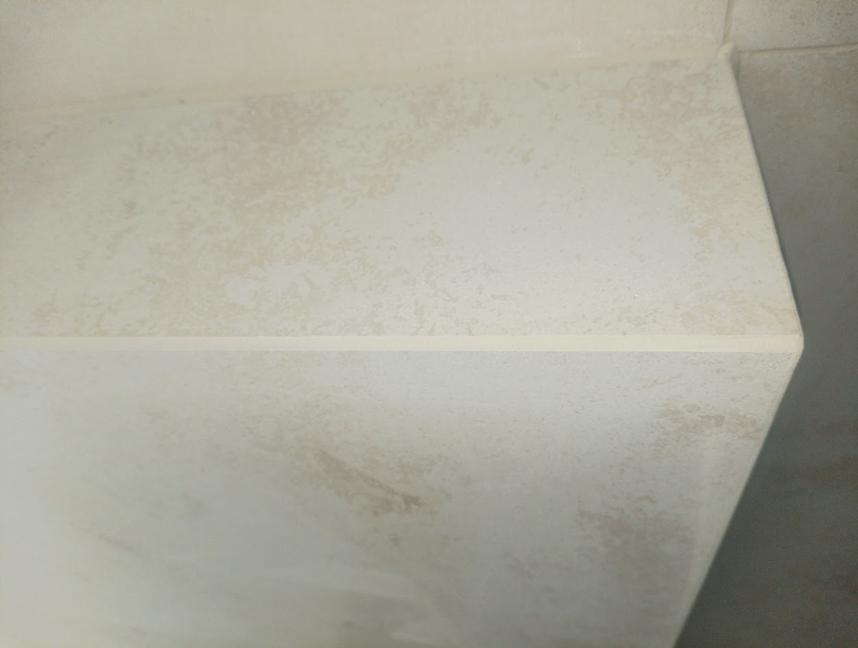 Jasna, beżowa ściana wykończona płytkami, z widoczną fugą. Płytki imitują naturalny kamień, tworząc subtelny wzór. Minimalistyczny detal wykończenia wnętrza.