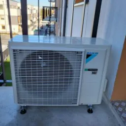 Biała jednostka zewnętrzna klimatyzatora Daikin zamontowana na balkonie nowego budownictwa, widok z góry na kratkę wentylacyjną i logo.