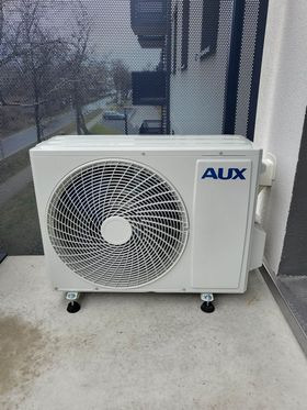Biała, zewnętrzna jednostka klimatyzacji marki AUX, zamontowana na balkonie z szarą posadzką, widok z góry, lekko z lewej strony.