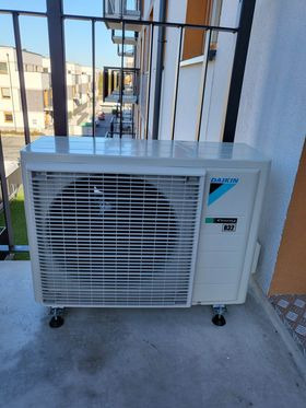 Biała jednostka zewnętrzna klimatyzatora Daikin zamontowana na balkonie nowego budownictwa, widok z góry na kratkę wentylacyjną i logo.