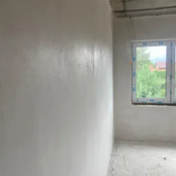 Świeżo położone, gładkie szare ściany w pokoju z surowym betonowym sufitem i oknem z widokiem na zieleń.