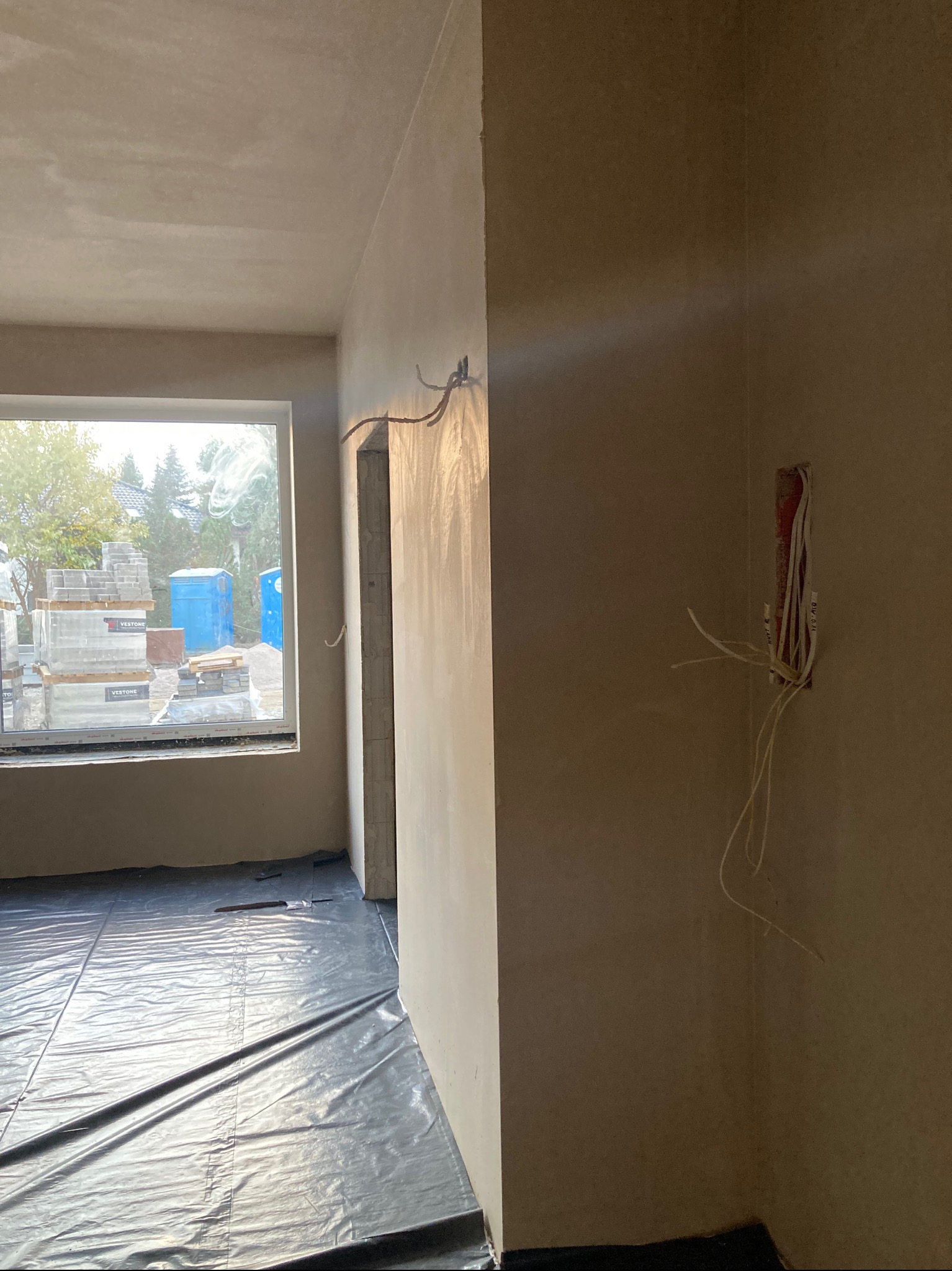 Wnętrze w trakcie prac wykończeniowych: świeżo otynkowane ściany, widoczne kable elektryczne i okno z widokiem na materiały budowlane Vestone oraz przenośną toaletę.