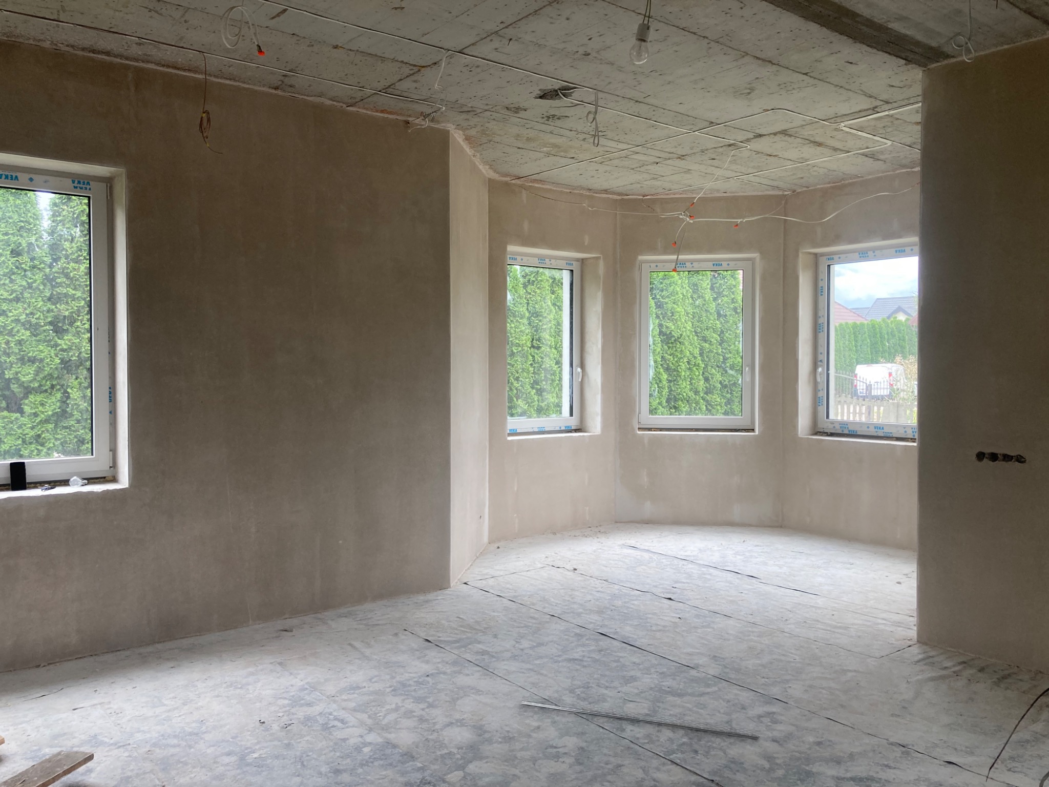Wnętrze surowego pomieszczenia po nałożeniu tynku na ściany, widoczne okna z zielonym krajobrazem na zewnątrz, betonowy sufit z instalacją elektryczną i podłoga przygotowana do dalszych prac...