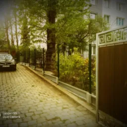 Nowoczesne ogrodzenie panelowe z zielonymi słupkami i betonową podmurówką, widoczne zza brązowej bramy wjazdowej na posesję z kostki brukowej, w tle zaparkowany samochód i budynek mieszkalny.