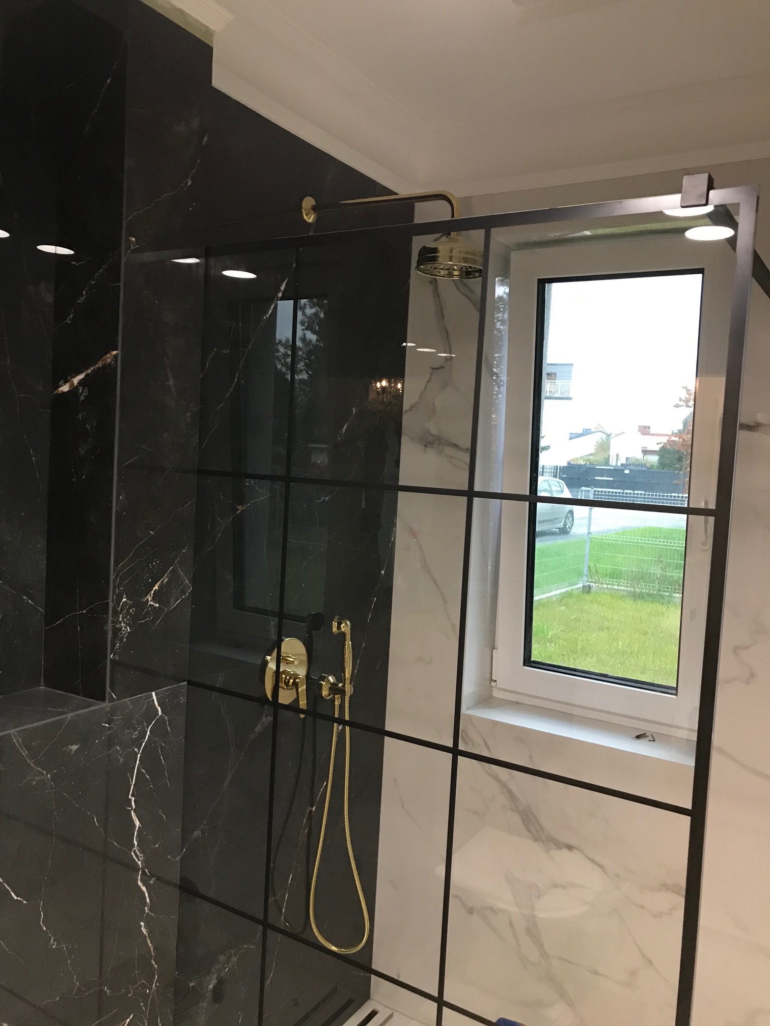 Nowoczesna łazienka z czarnymi marmurowymi płytkami z złotymi żyłami, kabina prysznicowa ze szklanymi panelami z czarnymi ramami, złota armatura i okno z widokiem na ogród.