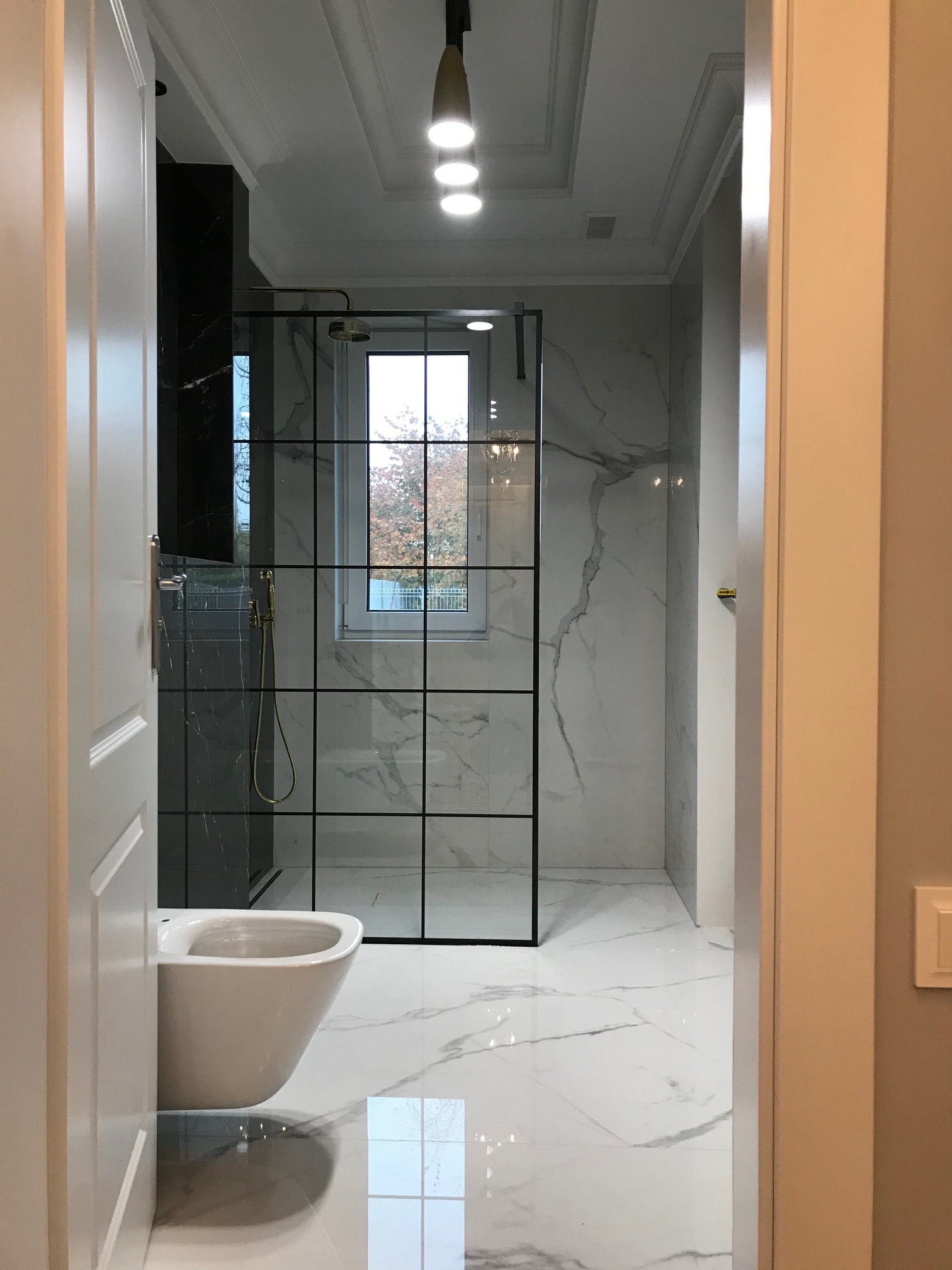 Nowoczesna łazienka z kabiną prysznicową w stylu loft, marmurowe płytki na ścianach i podłodze, wisząca miska WC, oświetlenie sufitowe punktowe, widok zza otwartych drzwi.