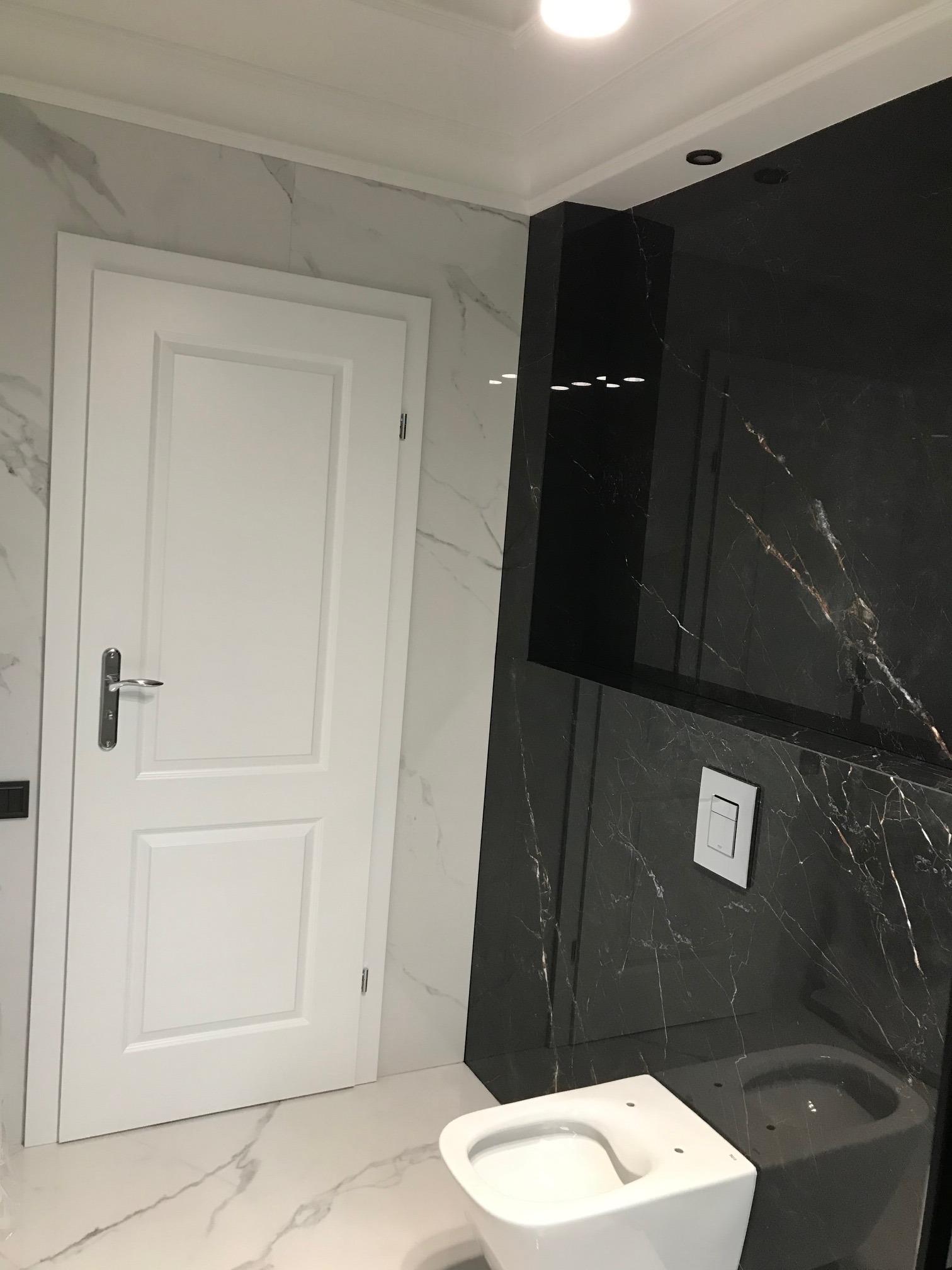 Nowoczesna łazienka z białymi marmurowymi płytkami na ścianach i podłodze, kontrastującymi z czarną ścianą z marmuru, biała wisząca toaleta i białe drzwi z chromowaną klamką.