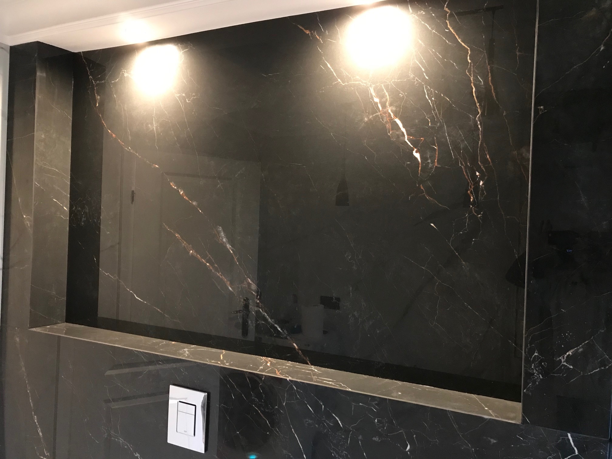 Narożna wnęka w ścianie wyłożona czarnym marmurem z białymi i złotymi żyłami, oświetlona dwoma punktowymi lampami, z widocznym włącznikiem światła na ścianie poniżej półki.