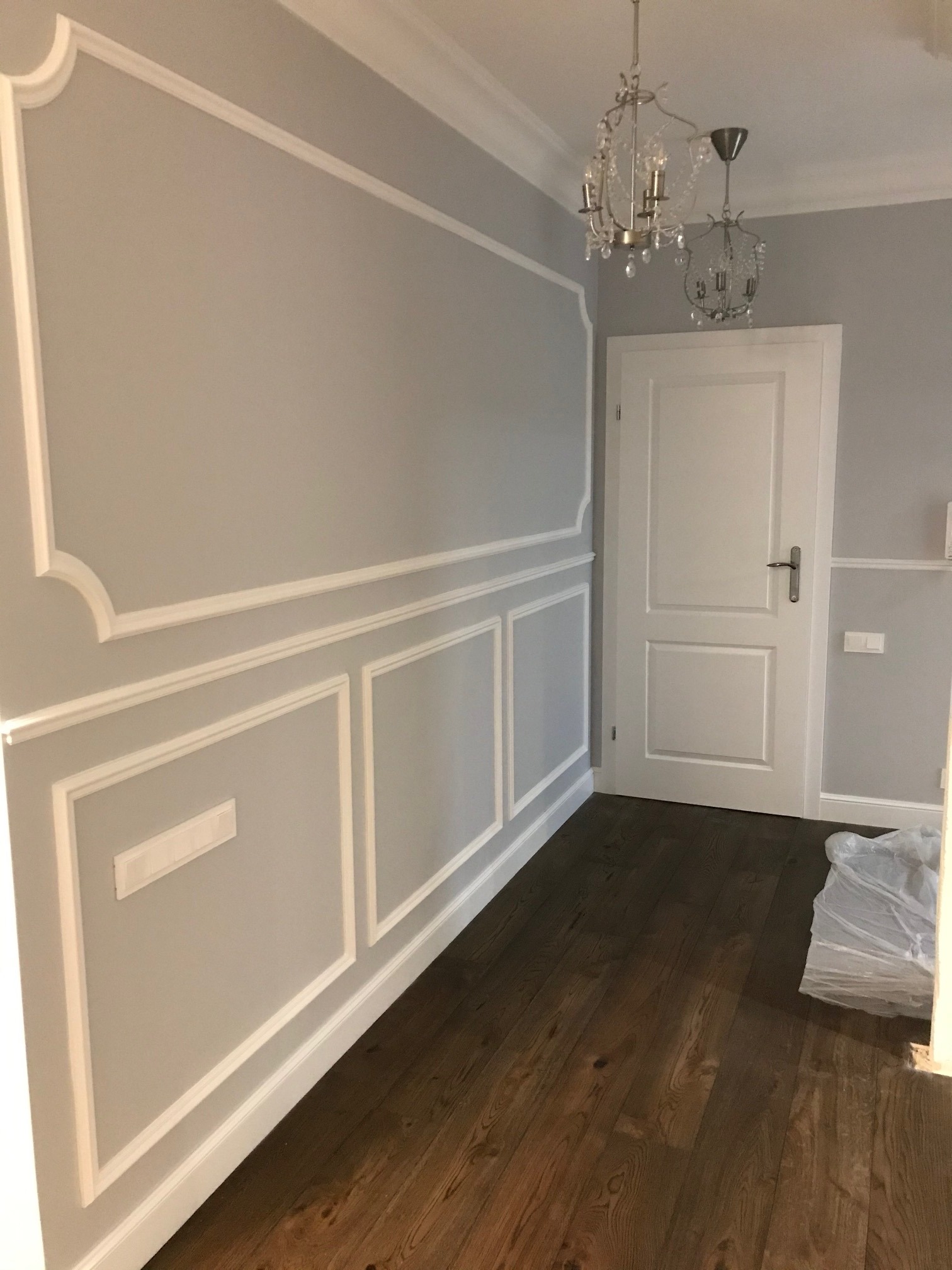 Szara ściana z dekoracyjnymi białymi listwami, drewniana podłoga, białe drzwi i kryształowe żyrandole.